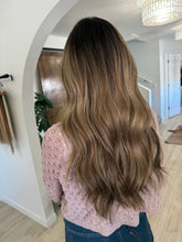 Load image into Gallery viewer, Person with long, wavy hair standing in a room with a mirror and light fixture.