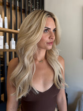Load image into Gallery viewer, Woman with long blonde hair in a room with shelves and bottles.
